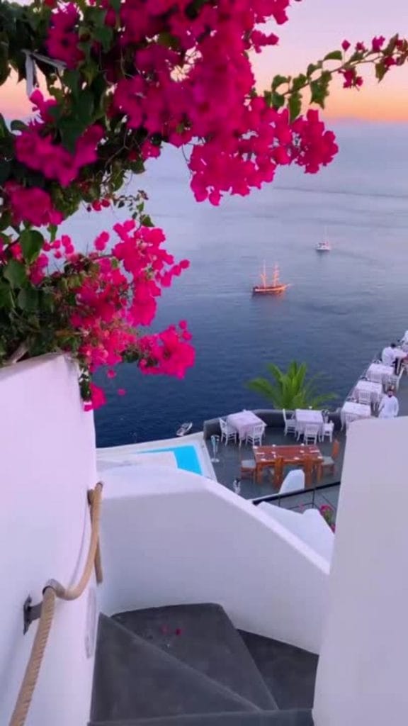 premium reel paket: Blick von einer weißen Treppe mit pinken Blumen auf das Meer bei Sonnenuntergang. Unten stehen weiße Tische und Stühle auf einer Terrasse mit Pool. Zwei Segelboote fahren auf dem ruhigen Wasser im Hintergrund.